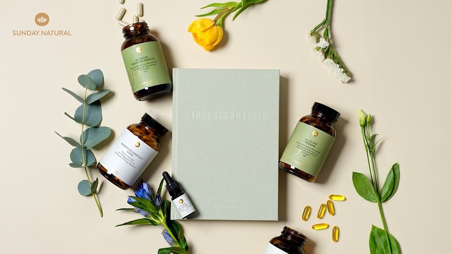Sunday Natural Flat Lay Aufnahme mit vier verschiedenen Nahrungsergänzungsmittelflaschen, einem True Clean Label Buch, Kapseln und frischen Pflanzenzweigen auf beigem Hintergrund.