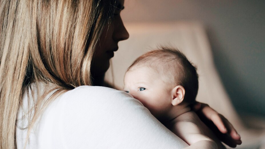 Nahaufnahme einer Mutter mit langen Haaren, die ihr Neugeborenes behutsam im Arm hält. Das Baby blickt über die Schulter der Mutter, während die Szene durch weiches, warmes Licht eine Atmosphäre von Geborgenheit und Ruhe vermittelt.