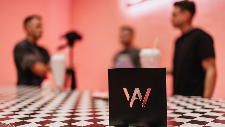 alt="Schwarzer Award-Aufsteller auf einer karierten Oberfläche vor pinkfarbenen Hintergrund mit Personen und Kamera-Equipment"