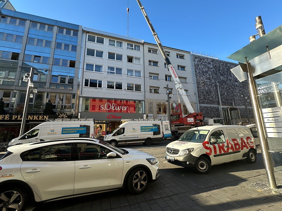 Kran hebt Material auf das Dach eines mehrstöckigen Gebäudes im Mannheimer Stadtzentrum; davor stehen Transporter von Biber Solarkonzept und ein STRABAG-Fahrzeug, Passantenbereich an den Planken.