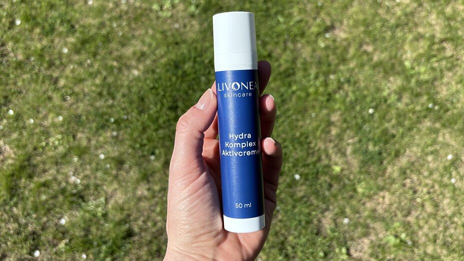 Eine Hand hält ein dunkelblaues 50-ml-Fläschchen der Marke LIVONEA Skincare mit der Aufschrift „Hydra Komplex Aktivcreme“ vor einem Hintergrund aus grüner Wiese.