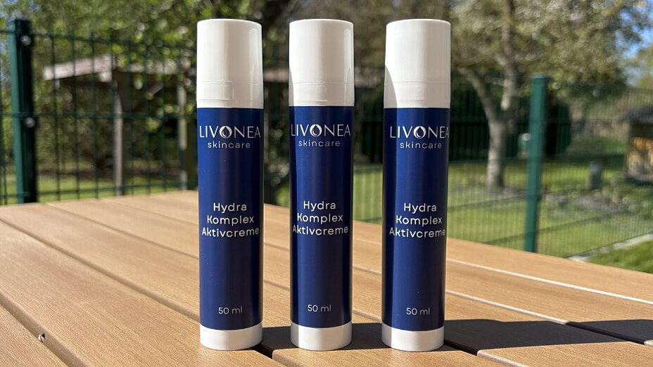 Drei dunkelblaue 50-ml-Spender der Marke LIVONEA Skincare mit der Aufschrift „Hydra Komplex Aktivcreme“ stehen auf einem hellbraunen Holztisch im Freien. Im Hintergrund ist ein grüner Garten mit Zaun erkennbar.