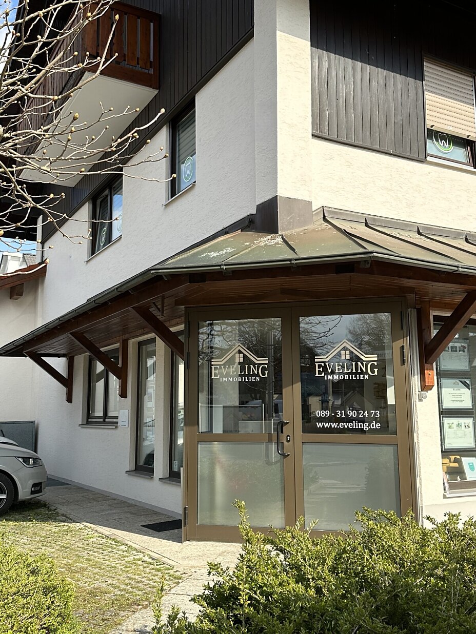 Bild von einer Gebäudeecke mit einer überdachten Glastür, auf der das Logo für Eveling Immobilien zu sehen ist. In Schaufenstern rechts und links der Tür befinden sich Immobilienaushänge.