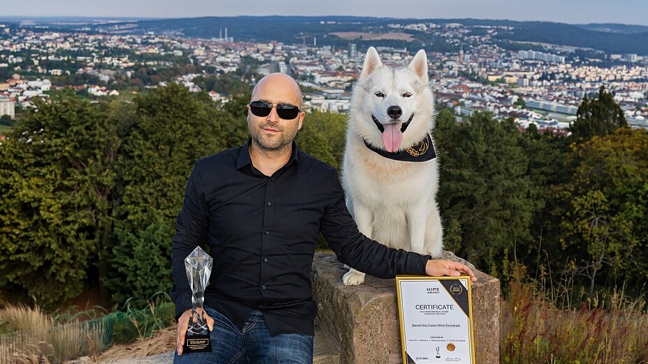 Special Dog Trainer Oliver Ehrentraut und sein weißer Husky Kuma posieren draußen glücklich mit dem HIPE-Award und der zugehörigen Auszeichnungsurkunde.