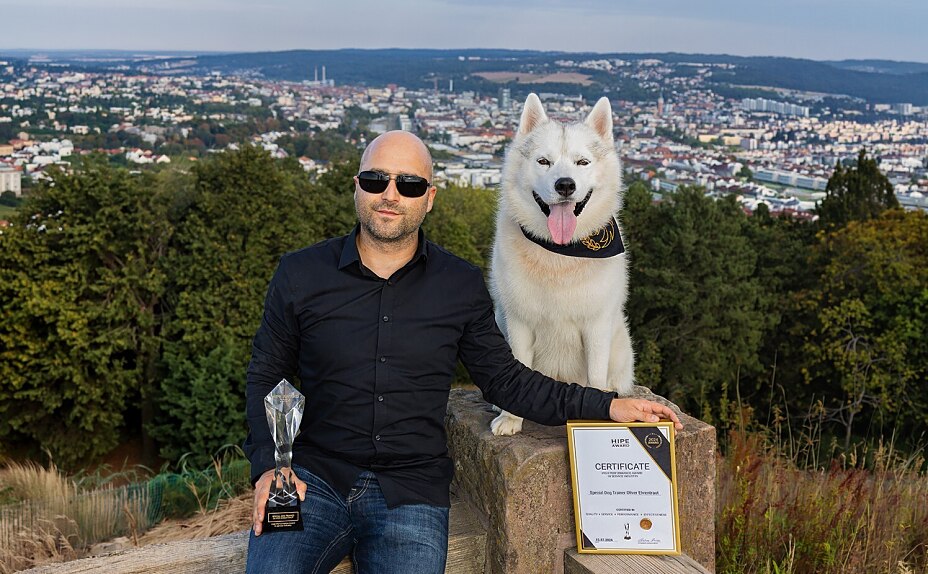 Special Dog Trainer Oliver Ehrentraut und sein weißer Husky Kuma posieren draußen glücklich mit dem HIPE-Award und der zugehörigen Auszeichnungsurkunde.